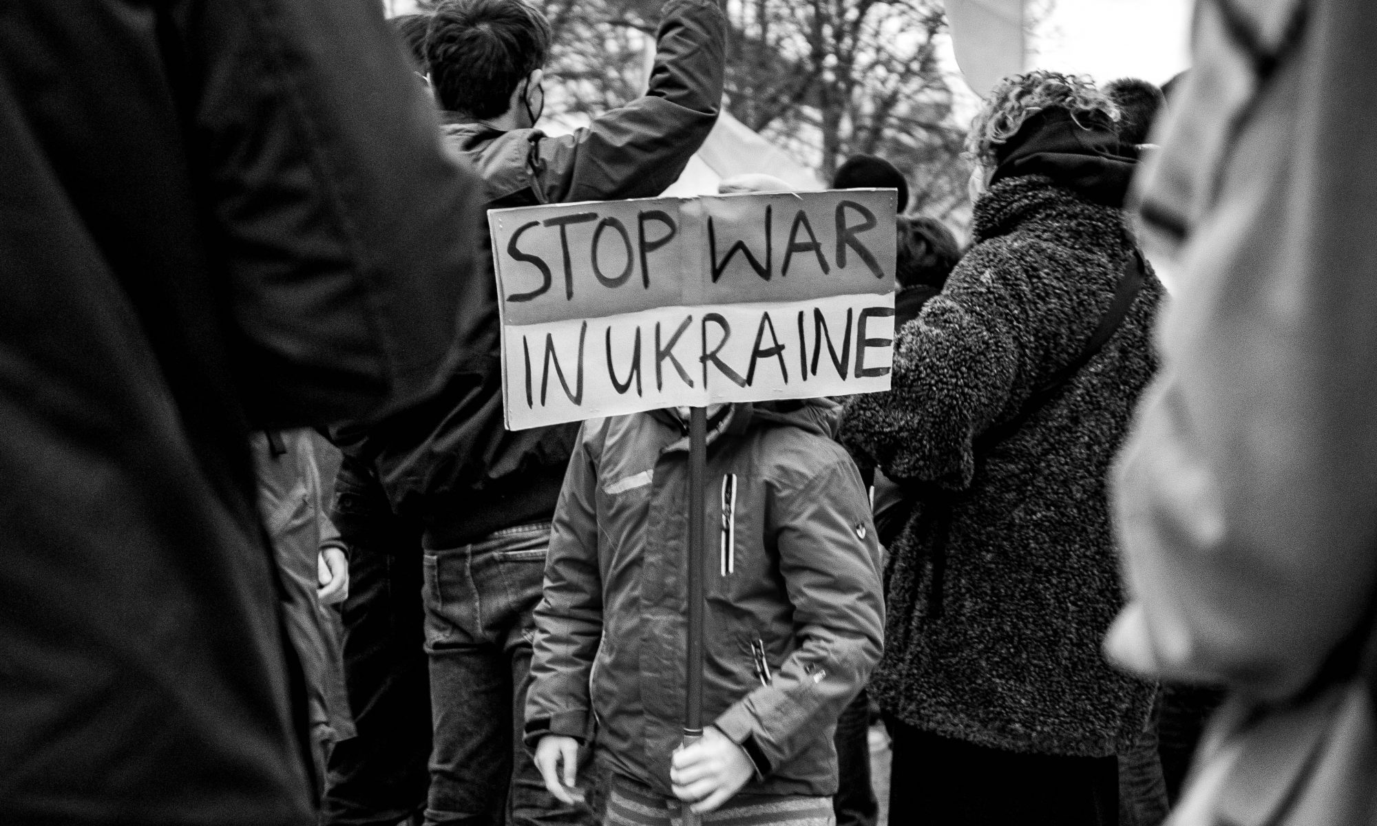 Kind hält ein Schild in der Hand: Stop war in Ukraine- Deutsch: Stoppt Krieg in der Ukraine. Das Gesicht des Kindes wird vom Schild verdeckt. Das Kind steht zwischen anderen Menschen. Das Photo ist schwarz-weiss