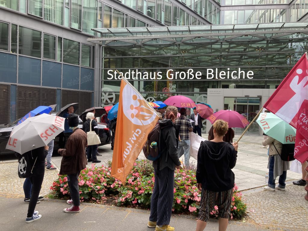 Das Bild zeigt die Demonstrationsgruppe von hinten vor dem Stadthaus Mainz. Eine Person trägt eine Fahne mit Logo von civi kune RLP