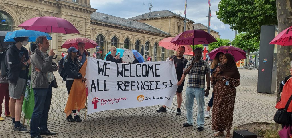 Auf dem Bild ist eine Gruppe von Menschen, vor dem Mainzer Hauptbahnhof mit Regenschirmen und einem Banner auf dem "We welcome all refugees" steht, auf deutsch "wir heißen alle Flüchtlinge willkommen". 