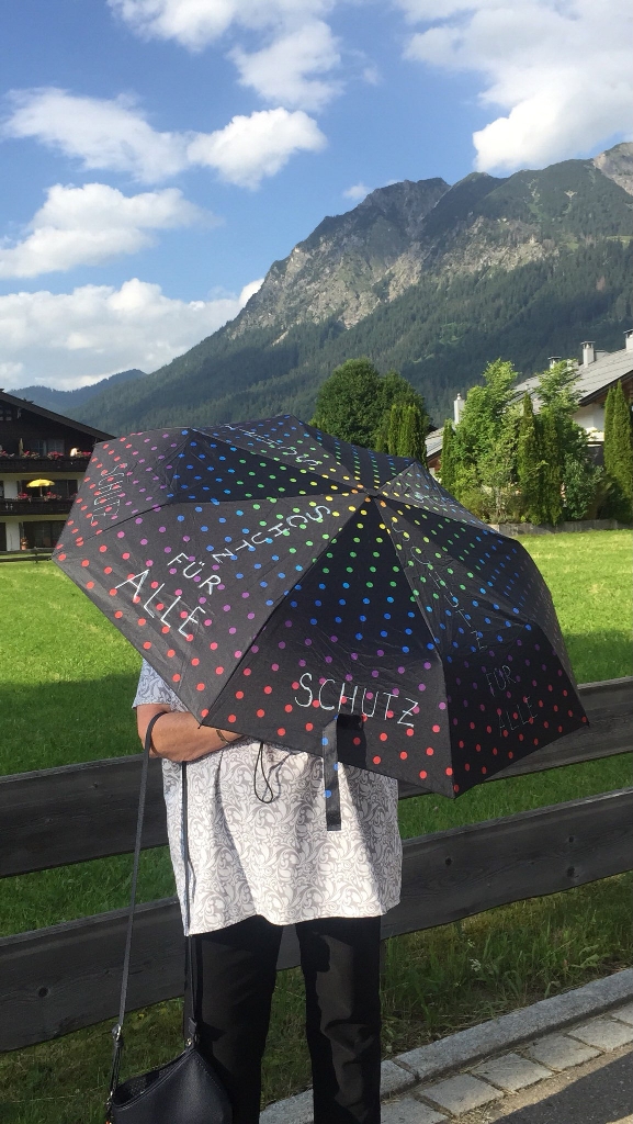 Eine Person hält einen Regenschirm vor sich. Auf dem Regenschirm steht:"Schutz für Alle". Im Hintergrund sieht man Berge. 