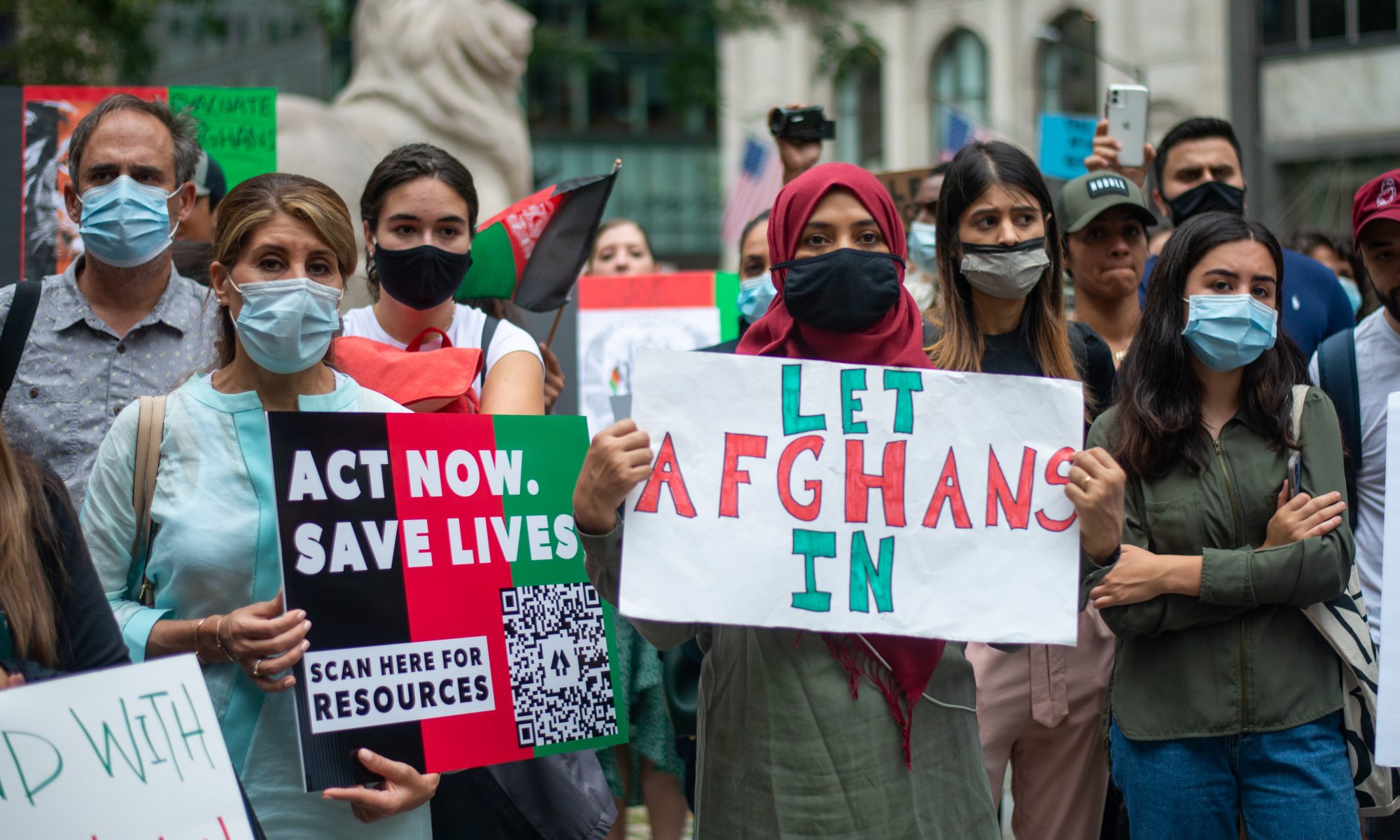 Eine Gruppe steht zusammen und hält Plakate hoch. Auf den Plakaten steht "Let Afghans in" und "Act now, save lifes"