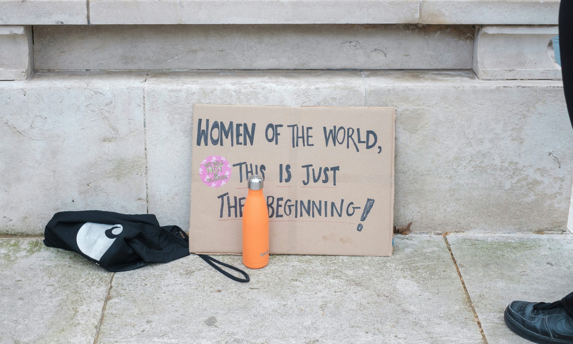 Plakatschild steht auf dem Boden. Drauf handschriftlich: "Women of the world, this is just the beginning! - auf detusch: Frauen der Welt, das ist erst der Anfang.